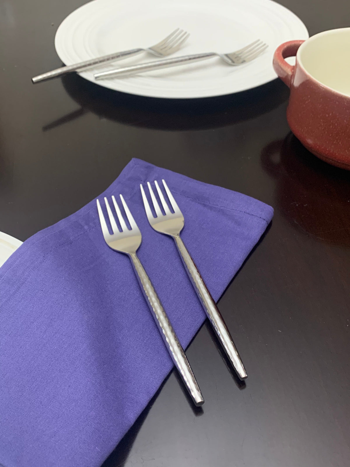 Two small stainless steel appetizer forks with textured handles resting on a folded purple cloth, with a white plate and fork in the background on a dark surface.