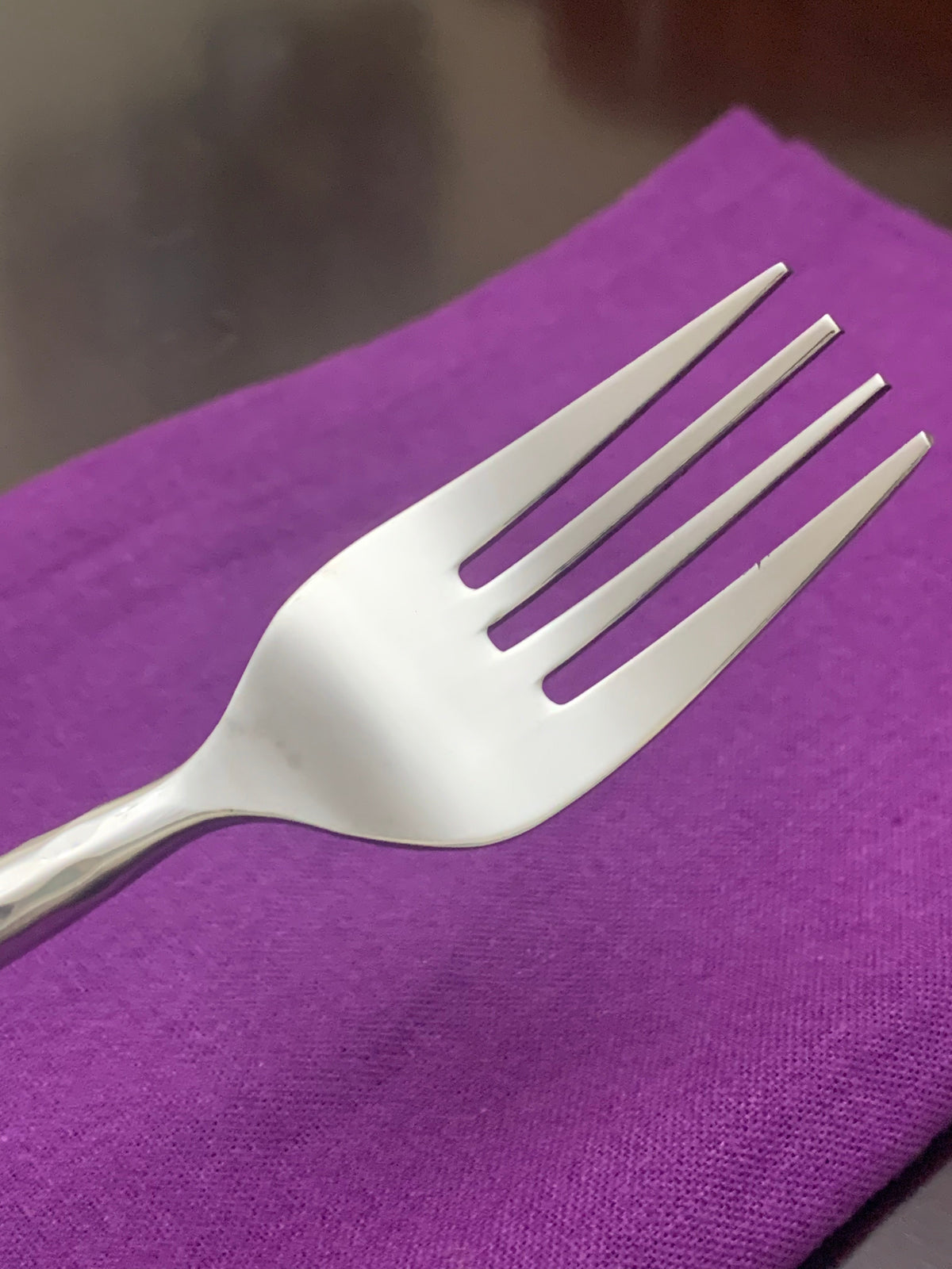 Close-up of a shiny stainless steel fork with four evenly spaced tines, resting on a vibrant purple cloth napkin over a dark brown surface.