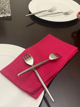 Two stainless steel appetizer forks with three prongs each, placed crossed on a folded red cloth napkin on a dark wood table near white plates and another pair of forks.