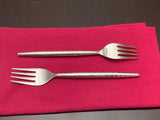 Two stainless steel salad appetizer forks with hammered handles, one placed horizontally and one vertically on a folded dark pink cloth against a dark surface.