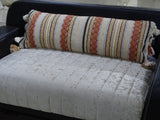 Long decorative accent throw pillow with horizontal textured stripes in beige, cream, rust orange, and black, featuring tassels at the ends, resting on a light quilted sofa seat.