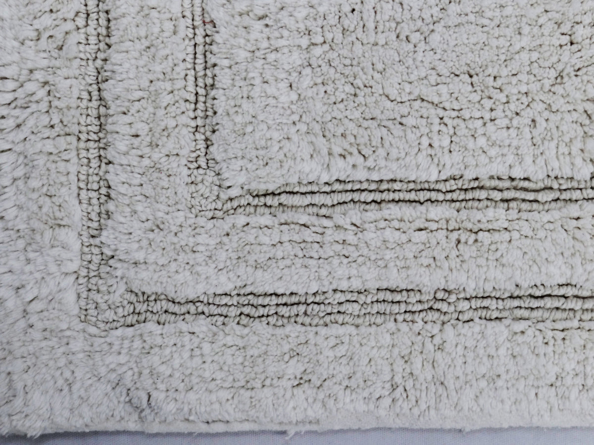 Close-up view of a plush ivory bath rug with a raised rectangular border pattern and a textured, fluffy surface.
