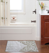 Beige and ivory bath rug with swirling damask pattern on a wooden floor in front of a white bathtub and brown wooden vanity with flowers.