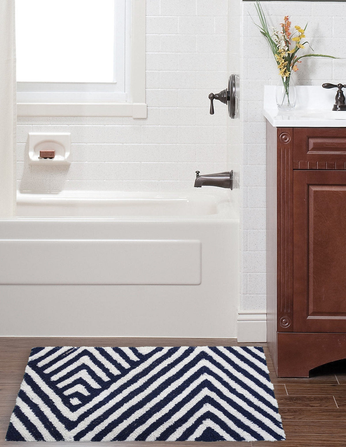 Rectangular bath rug with a charcoal and ivory chevron pattern placed on dark wood floor next to a brown vanity and white tiled bathtub area.