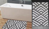 Rectangular grey and ivory bath rug with interlocking geometric angled line patterns, placed on wood-look floor next to a white, rectangular bathtub with silver fixtures.