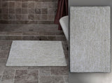 Rectangular ivory and white bath rug with a simple textured pattern placed on gray stone tile bathroom floor next to a white bathtub with a burgundy curtain.
