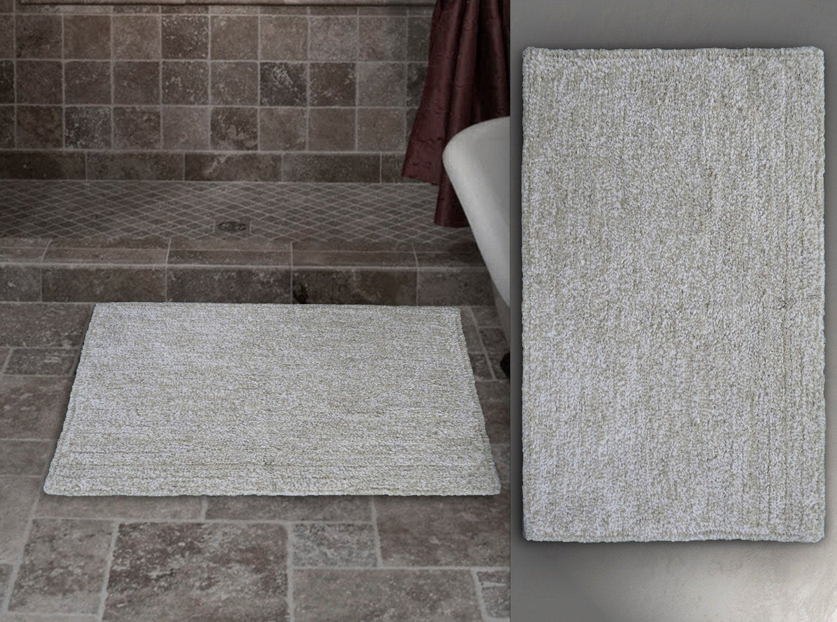 Rectangular ivory and white bath rug with a simple textured pattern placed on gray stone tile bathroom floor next to a white bathtub with a burgundy curtain.