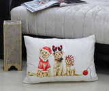 Rectangular white throw pillow with illustration of three kittens dressed in Christmas attire, including a Santa hat, reindeer antlers, and striped elf hat, placed on floor near beige upholstered couch.
