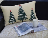 Rectangular beige pillow with three green Christmas trees topped with red ornaments, placed on a beige cushioned surface next to an open magazine and white ceramic mug.