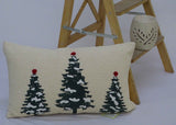 Rectangular beige throw pillow featuring three embroidered dark green Christmas trees with scattered white snow and red pom-pom toppers, set against a plain background with a wooden ladder and decorative candle holder.