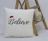 Ivory textured cushion with the word 'Believe' embroidered in black and a small red and white Santa hat detail on the first letter, placed against a wooden ladder with a carved candle holder.