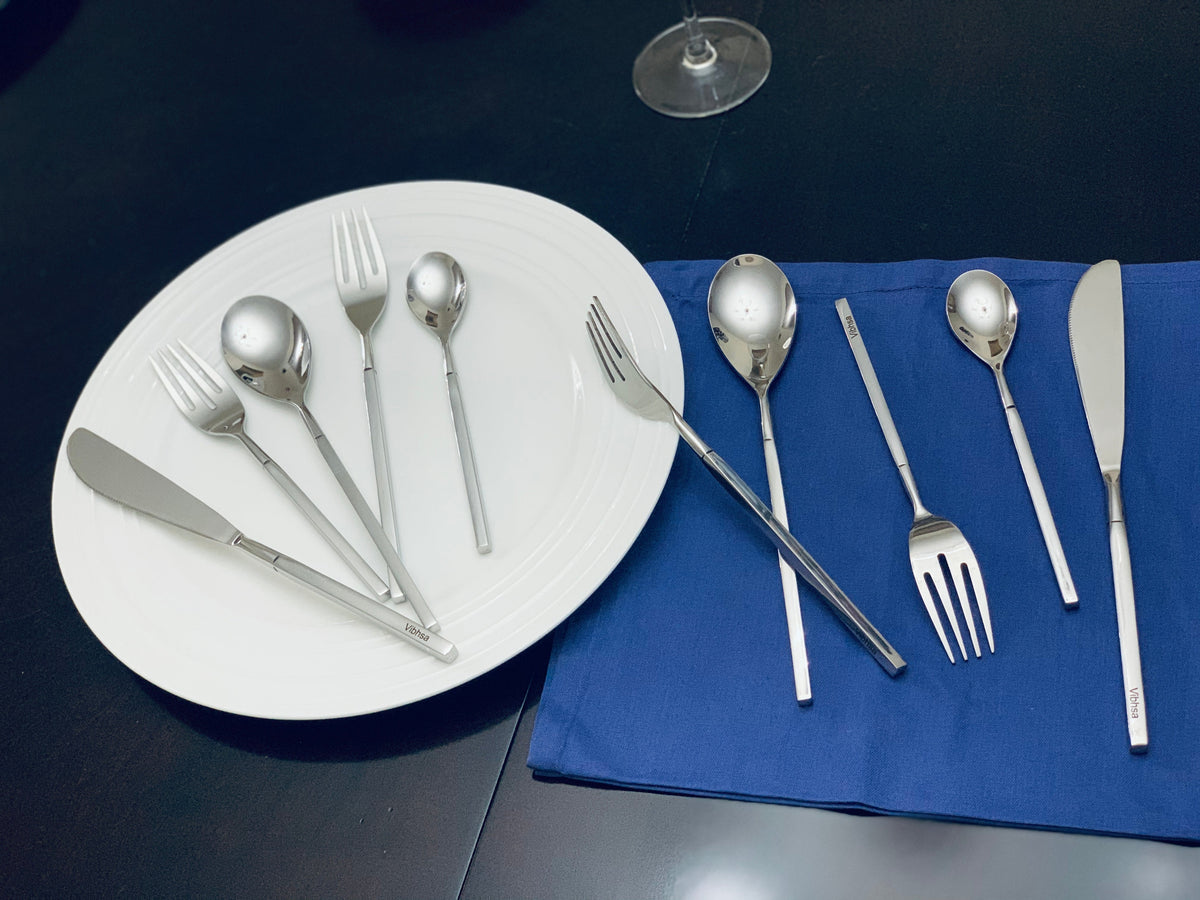 Modern stainless steel flatware pieces including forks, knives, and spoons arranged on a white plate and a blue cloth on a black table.
