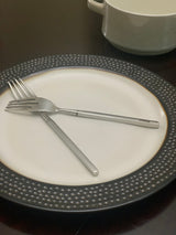 Two modern stainless steel forks with flat handles resting crossed on a white plate with a textured dark gray rim, a white ceramic pot in background