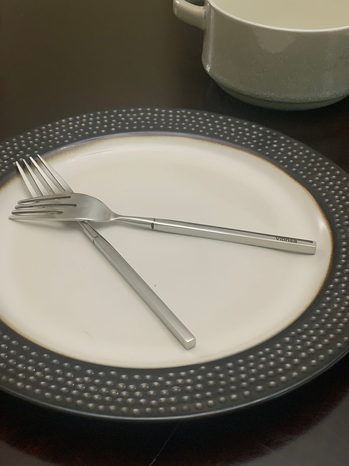 Two modern stainless steel forks with flat handles resting crossed on a white plate with a textured dark gray rim, a white ceramic pot in background