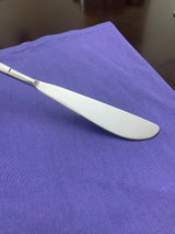 Silver stainless steel butter knife with a rounded, slightly serrated blade resting on a textured purple cloth against a dark brown surface.