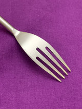 Close-up of a stainless steel fork with four tines against a purple fabric background, highlighting the flatware's smooth, modern design and matte finish.