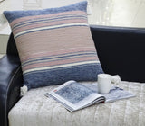 Square 22-inch throw pillow with horizontal blue, beige, and pink stripes and white tassels on dark couch, next to open magazine and white mug on light quilted fabric.