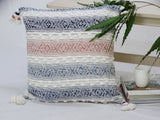 Square throw pillow with blue, pink, and white horizontal woven stripes and tassels on each corner, placed on a white surface near a wooden chair and books.