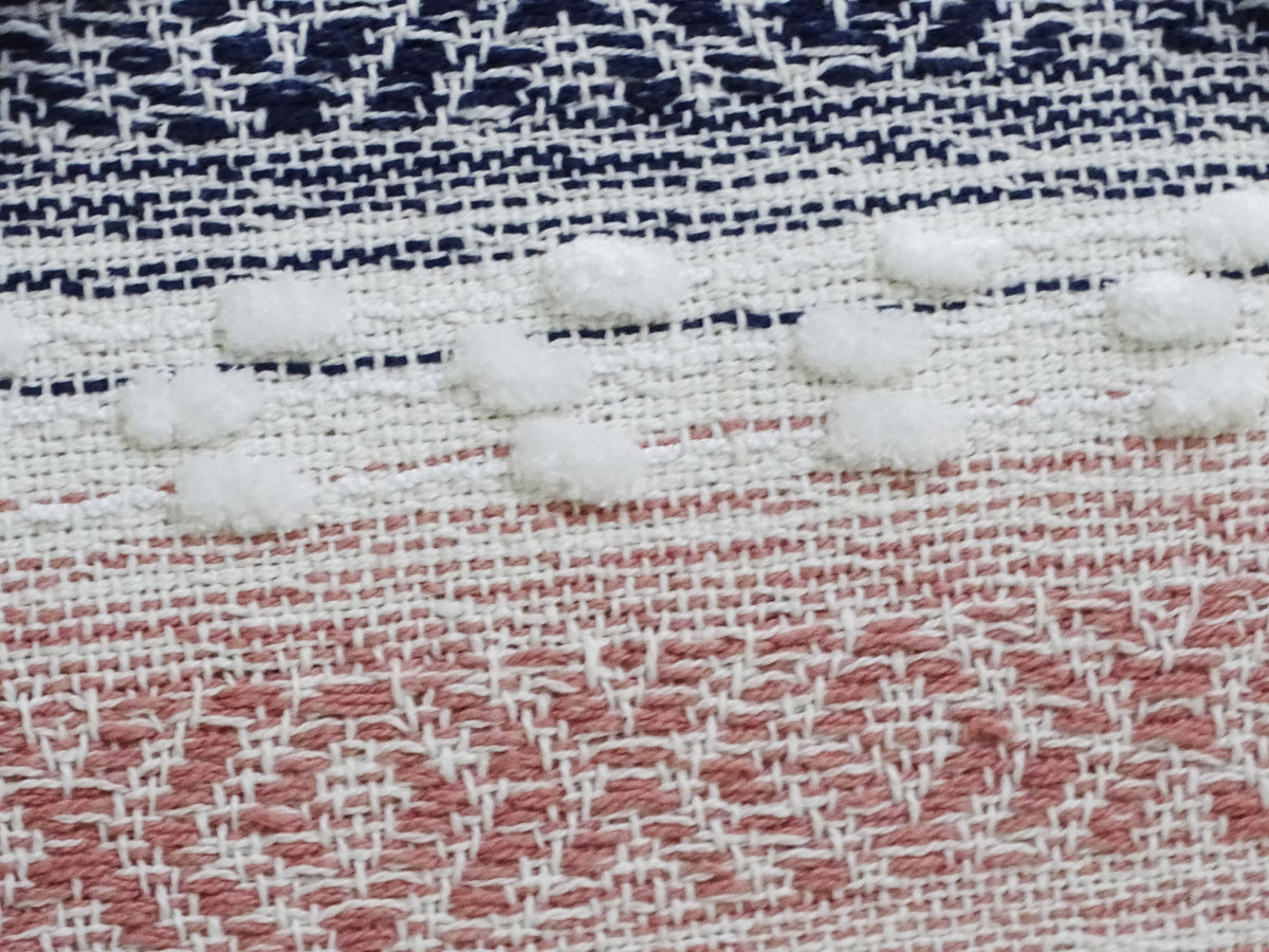 Close-up of a woven textile showing white textured dots over navy blue and white thread, and a dusty pink and white geometric pattern below.
