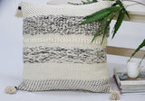 Square throw pillow with textured woven stripes in cream, black, and beige, featuring tassels on each corner, beside a light wooden table with a green leafy plant and stacked books.