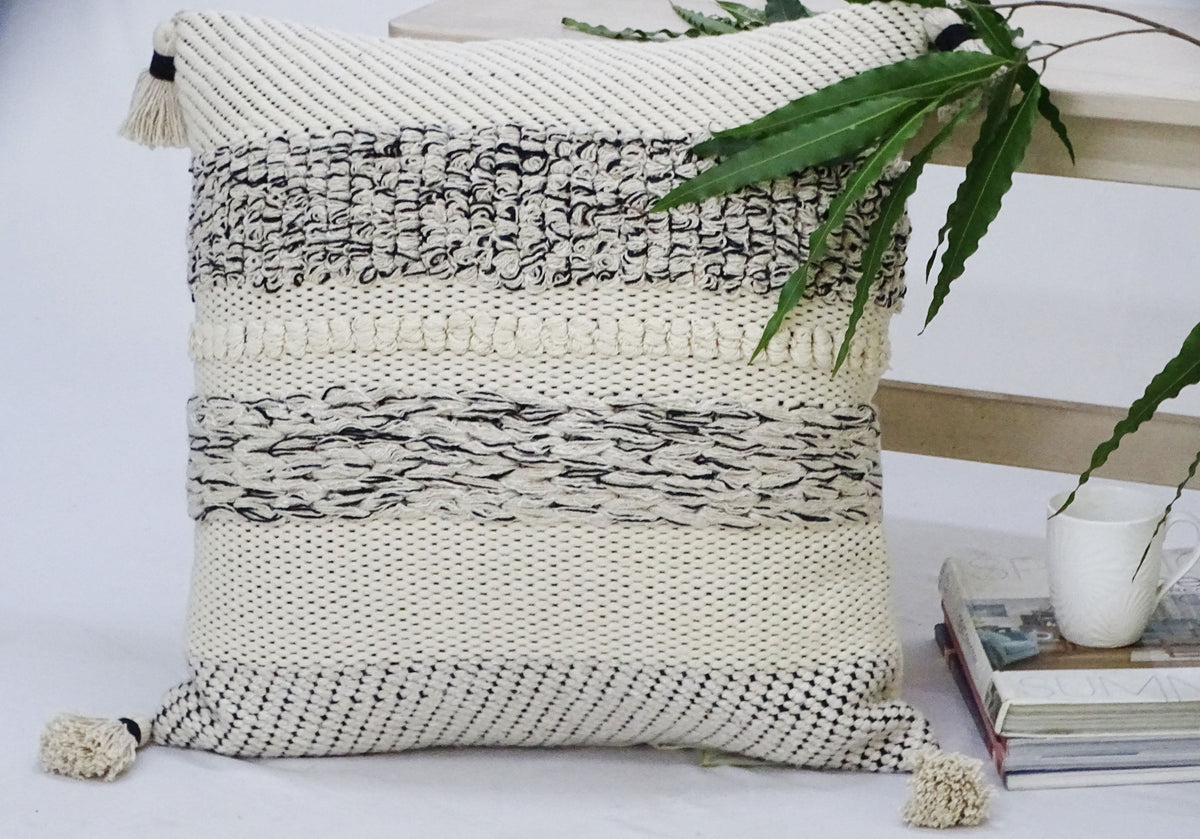 Square throw pillow with textured woven stripes in cream, black, and beige, featuring tassels on each corner, beside a light wooden table with a green leafy plant and stacked books.