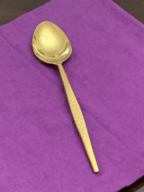 Single gold spoon with a hammered texture handle placed diagonally on a purple fabric background.