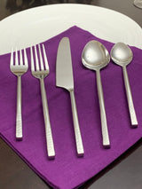 Five-piece silver flatware set including two forks, a knife, a large spoon, and a small spoon, arranged on a purple napkin on a white plate.