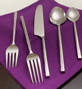 Five silver flatware pieces with hammered texture handles including two forks, a butter knife, and two spoons on a purple cloth.