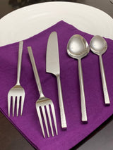 Five-piece silver flatware set with hammered handles including two forks, a butter knife, a large spoon, and a small spoon arranged on a folded purple napkin.