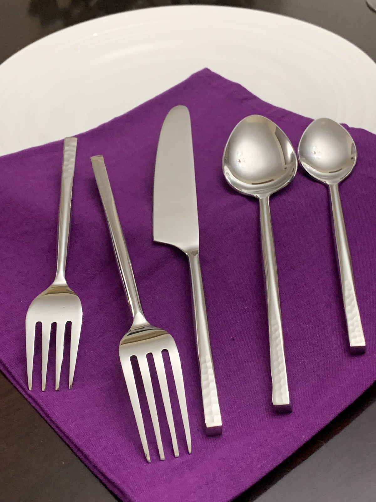 Five-piece silver flatware set with hammered handles including two forks, a butter knife, a large spoon, and a small spoon arranged on a folded purple napkin.