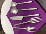 Five silver flatware pieces including two ladles, a butter knife, a dinner fork, and a salad fork with hammered handles on a purple cloth over a large white plate.