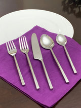 Five-piece silver flatware set including two forks, a knife, a tablespoon, and a teaspoon, arranged on a purple cloth with a white plate in the background.