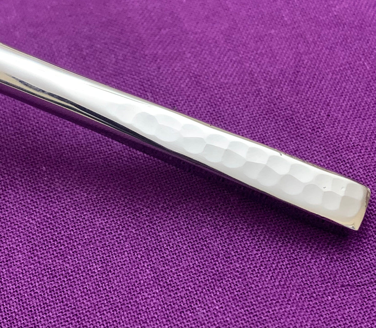 Close-up of a silver flatware handle with a hammered texture against a purple fabric background, highlighting the subtle dimples and smooth polish.