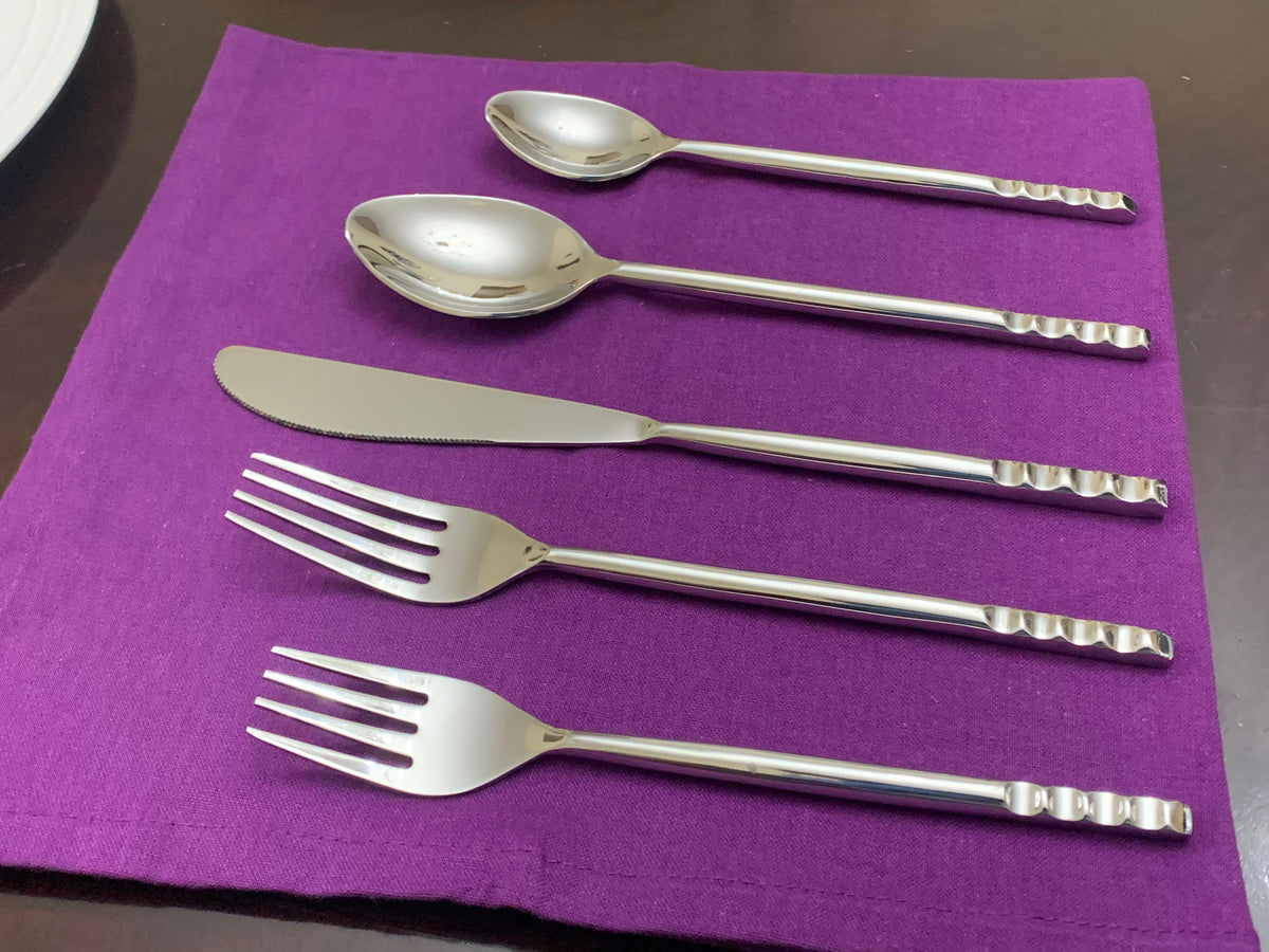 Five-piece stainless steel flatware set including two spoons, one knife, and two forks with textured handles, arranged on a purple cloth on a dark table.