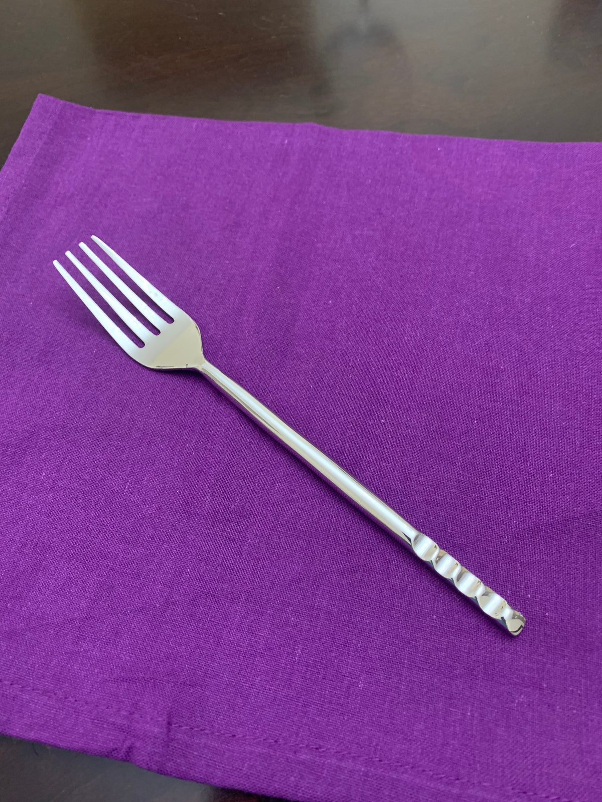 Silver fork with a modern, ribbed handle design resting on a purple cloth napkin over a dark wooden surface.