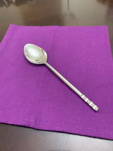 Shiny silver spoon with a distinctive beaded handle resting on a purple cloth atop a dark wooden surface.