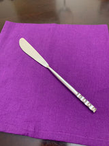 Silver butter knife with a smooth blade and a handle featuring a textured, grooved pattern, placed on a folded purple cloth napkin on a wooden surface.