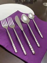 Five-piece stainless steel flatware set including two forks, a butter knife, and two spoons, arranged on a purple cloth napkin atop a dark wood table