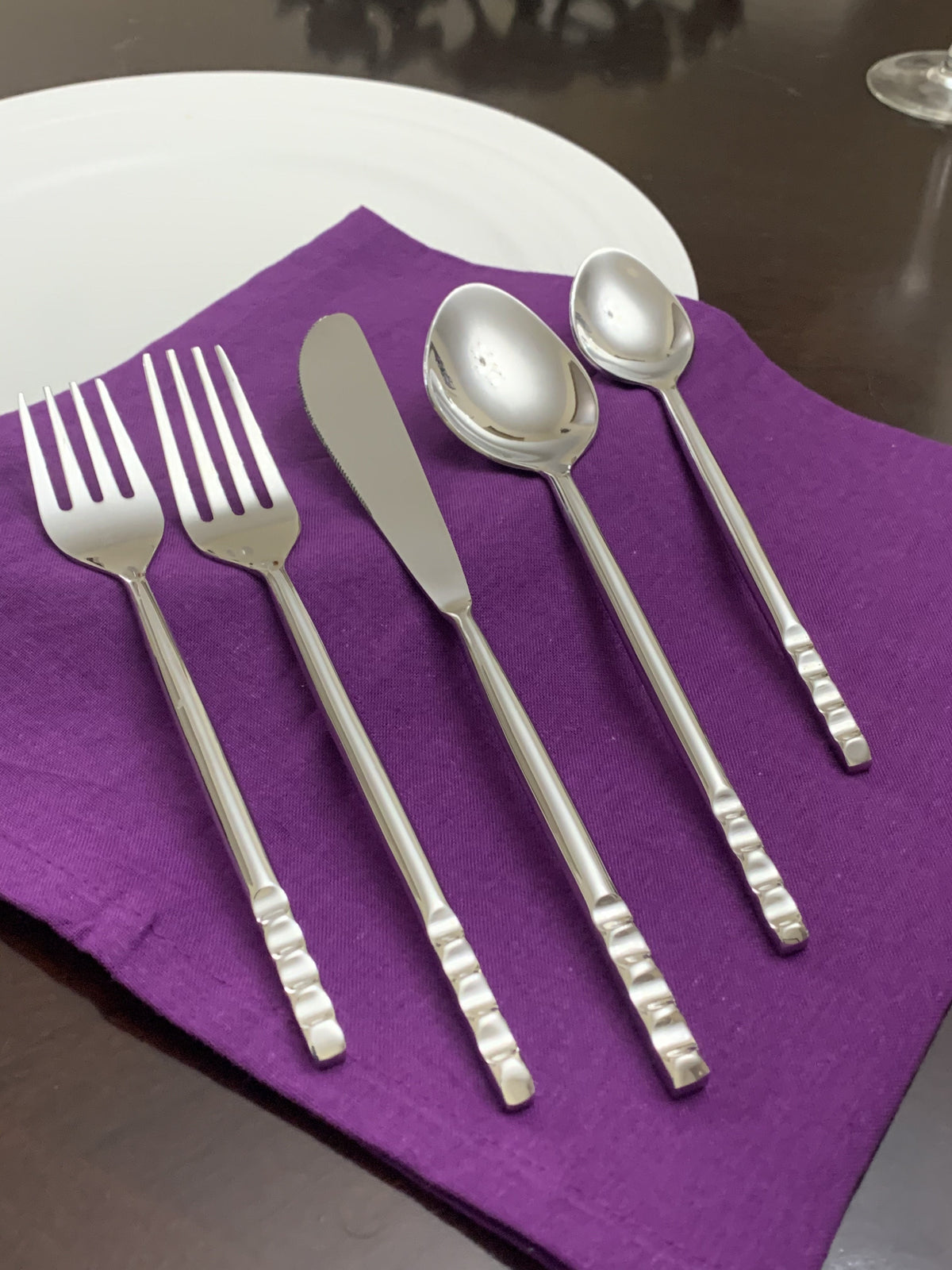 Five-piece stainless steel flatware set including two forks, a butter knife, and two spoons, arranged on a purple cloth napkin atop a dark wood table