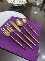 Five-piece gold flatware set including two forks, a knife, and two spoons laid on a purple cloth napkin with a white plate and wine glass in the background