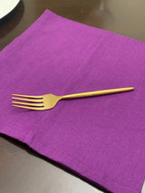 Single golden fork with a sleek, modern design resting on a vibrant purple cloth napkin atop a dark wooden table.