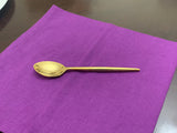 Single golden teaspoon with smooth polished finish placed centrally on a purple fabric square on a dark wood surface