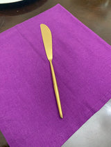 Single gold-toned butter knife with a smooth, rounded blade resting on a bright purple cloth napkin on a dark wooden surface.
