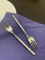 Two stainless steel dinner forks with straight handles inscribed 'Vibhsa', placed on a purple cloth next to a white plate on a dark surface.