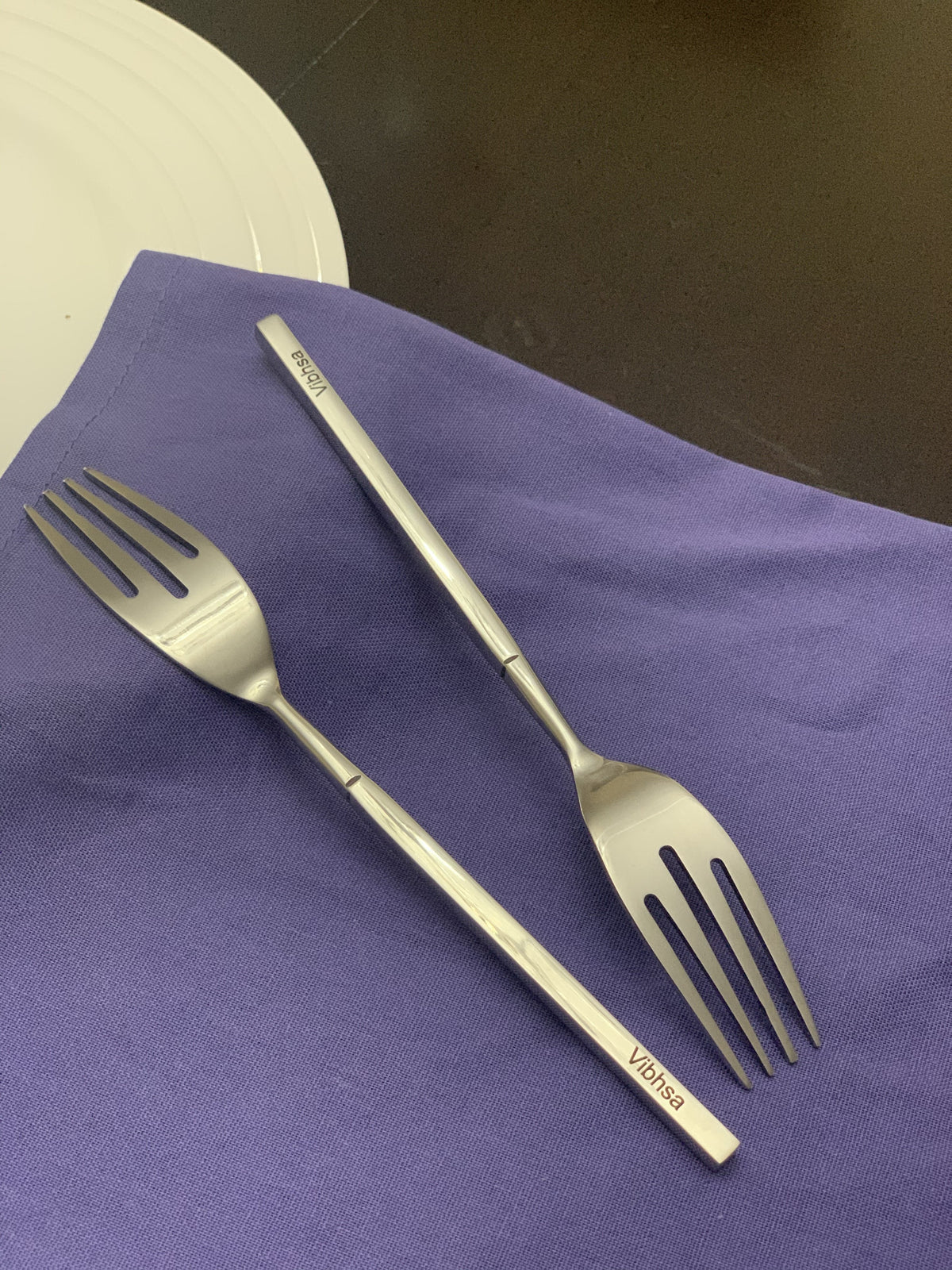 Two stainless steel dinner forks with straight handles inscribed 'Vibhsa', placed on a purple cloth next to a white plate on a dark surface.