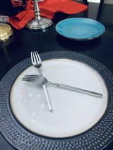 Two stainless steel dinner forks with slim handles crossed on a white plate with a textured black rim, against a table setting with a blue ceramic plate in the background.