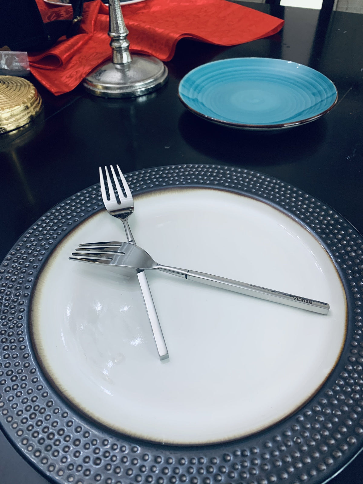 Two stainless steel dinner forks with slim handles crossed on a white plate with a textured black rim, against a table setting with a blue ceramic plate in the background.
