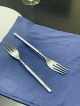 Two stainless steel dinner forks with four tines and straight handles, one showing the brand name 'Vibhsa,' placed on a blue cloth on a dark surface.