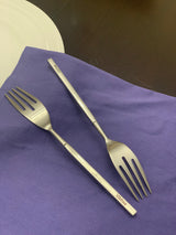 Two stainless steel salad forks with a matte finish and three wide tines, placed on a purple cloth next to a white plate on a dark surface.