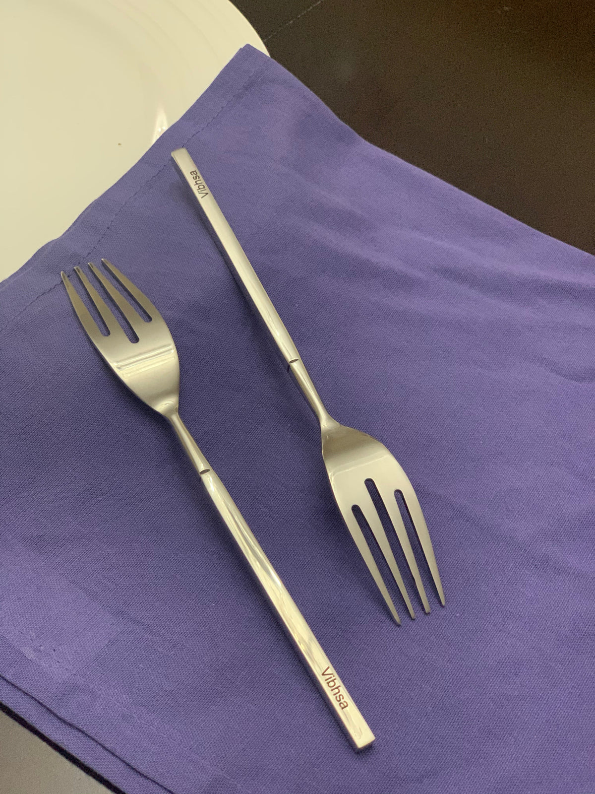 Two stainless steel salad forks with slender, slightly curved handles, one showing the engraved brand name 'Vibhsa,' placed on a purple cloth against a dark surface.